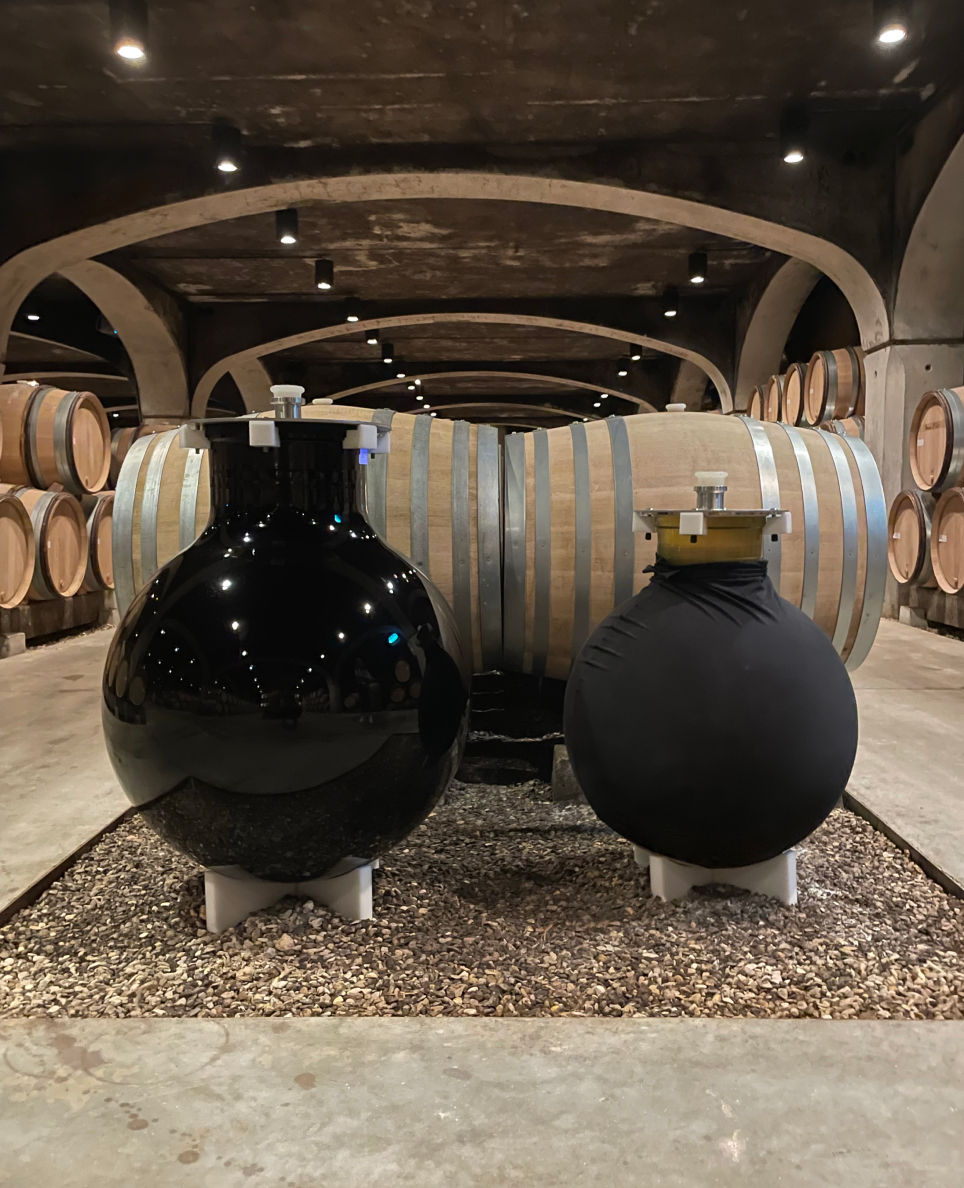 deux cuves en verres wineglobe masquées par un tissu noir, dans un caveau avec des tonneaux en bois@2x